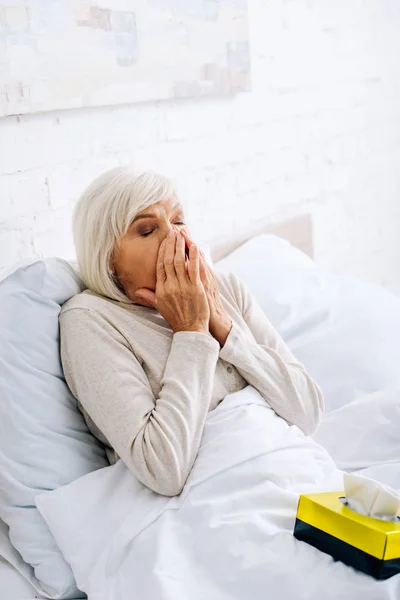 Attractive and ill senior woman with closed eyes sneezing in bed — Stock Photo