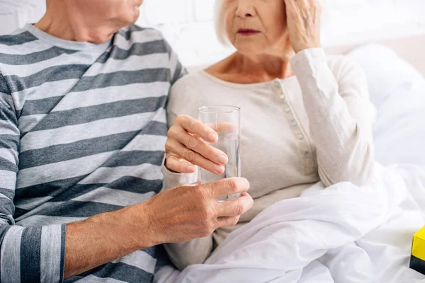 Vista recortada del marido dando vaso de agua a la esposa con dolor de cabeza en el apartamento - foto de stock