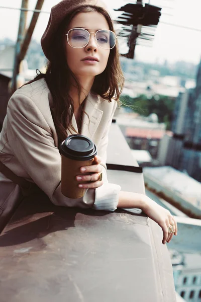 Привлекательная элегантная девушка в бежевом костюме и берете позирует на крыше с кофе, чтобы пойти — стоковое фото