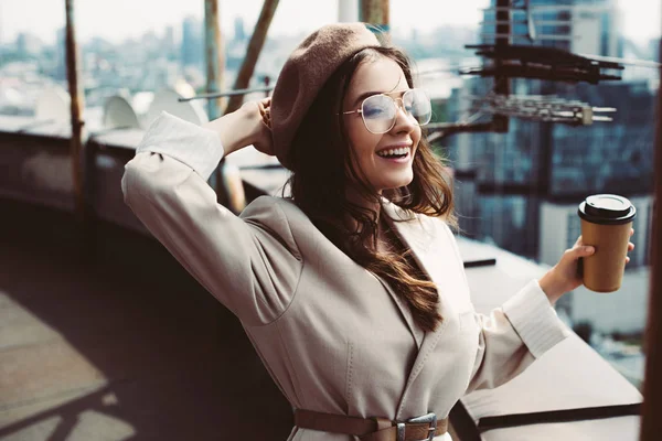 Atractiva chica feliz en traje beige y boina posando en el techo con café para llevar - foto de stock