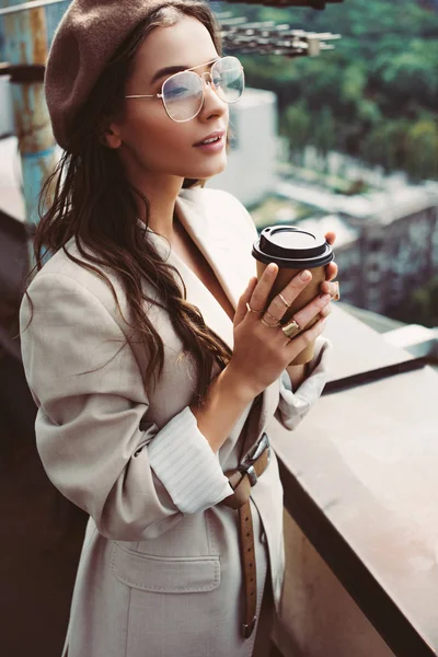 Atractiva chica de moda en traje beige y boina posando en el techo con café para llevar - foto de stock