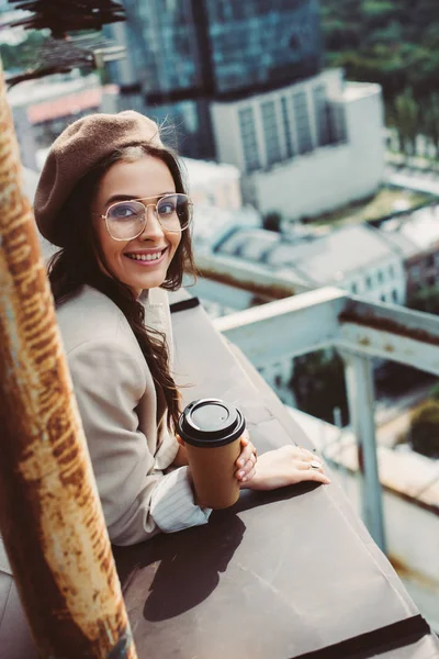 Souriant fille à la mode en costume beige et béret posant sur le toit avec du café à emporter — Photo de stock
