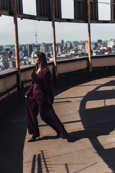 Attractive elegant woman posing in trendy burgundy suit and sunglasses on urban roof — Stock Photo