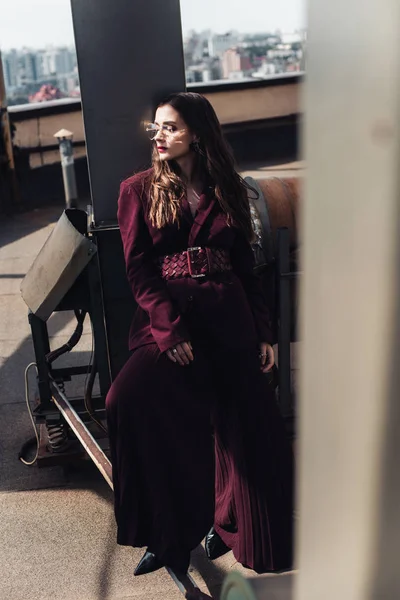 Femme élégante posant en costume bordeaux tendance et lunettes de soleil sur le toit urbain — Photo de stock