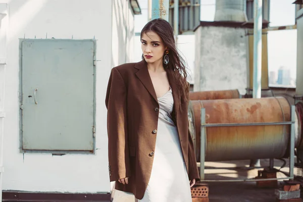 Elegante chica de moda posando en vestido de seda y chaqueta marrón en el techo - foto de stock