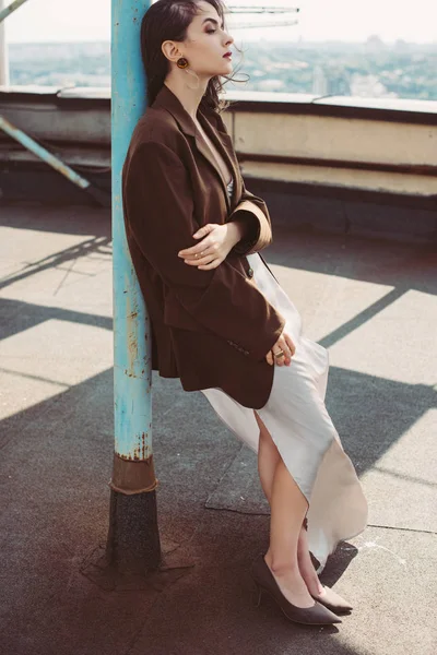 Hermosa chica de moda posando en vestido de seda y chaqueta marrón en el techo - foto de stock