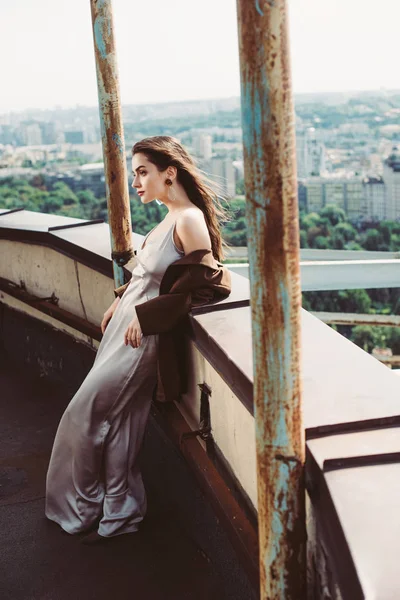 Chica elegante de moda posando en vestido de seda y chaqueta marrón en el techo urbano - foto de stock