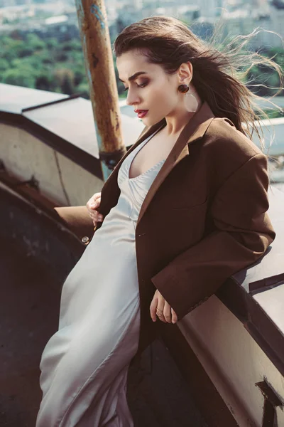 Stylish elegant girl posing in silk dress and brown jacket on roof — Stock Photo