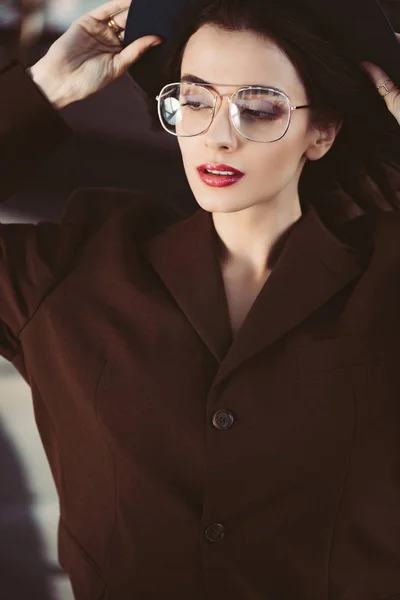 Elegant trendy woman posing in hat, eyeglasses and brown jacket on roof — Stock Photo