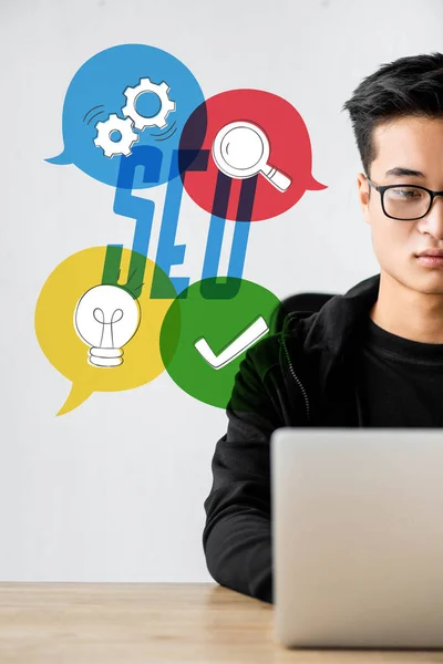 Cropped view of asian seo manager using laptop and sitting near illustration with seo lettering — Stock Photo