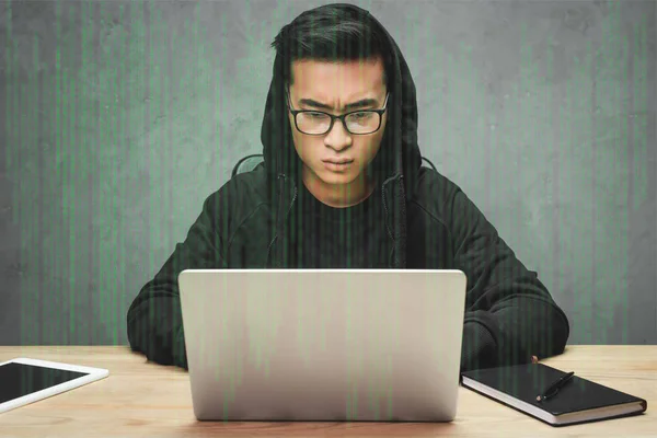 Asian hacker in glasses sitting at table, using laptop and surrounded with illustration — Stock Photo