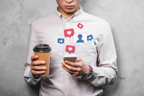 Vista recortada del gerente seo sosteniendo la taza de papel, utilizando el teléfono inteligente con ilustración emojis - foto de stock