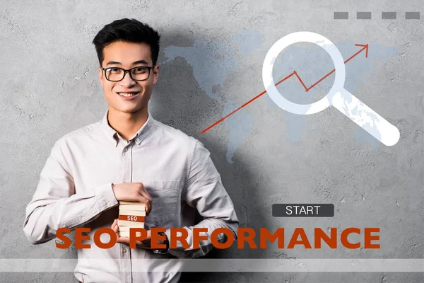 Smiling asian seo manager holding rectangle with seo lettering and standing near seo performance illustration — Stock Photo