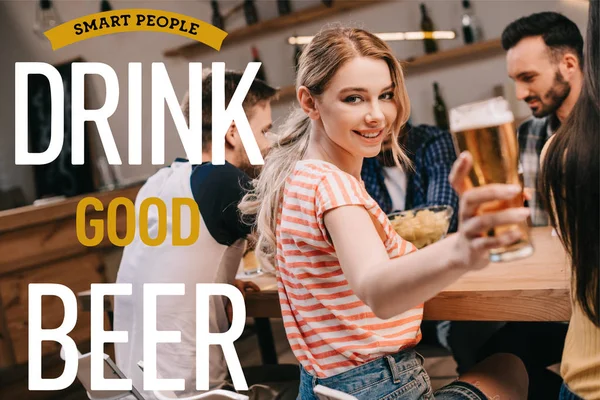 Selective focus of smiling young woman looking at camera while holding glass of light beer near smart people drink good beer illustration — Stock Photo