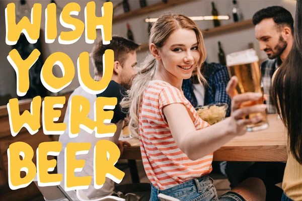 Selective focus of smiling young woman looking at camera while holding glass of light beer near wish you were beer illustration — Stock Photo