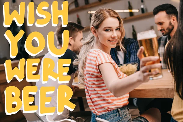 Foyer sélectif de sourire jeune femme regardant la caméra tout en tenant un verre de bière légère près de souhaiter que vous étiez illustration de bière — Photo de stock