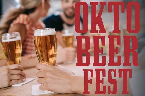 Cropped view of women holding glasses of light beer while sitting together with friends in pub near oktobeerfest illustration — Stock Photo