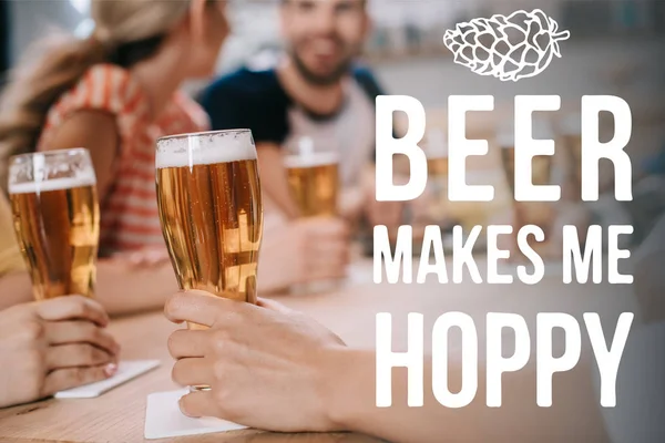 Cropped view of women holding glasses of light beer while sitting together with friends in pub near beer makes me hoppy illustration — Stock Photo