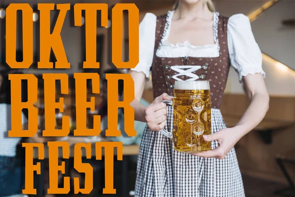 Vista parcial de camarera en traje nacional alemán sosteniendo taza de cerveza ligera cerca de Oktobeerfest ilustración - foto de stock