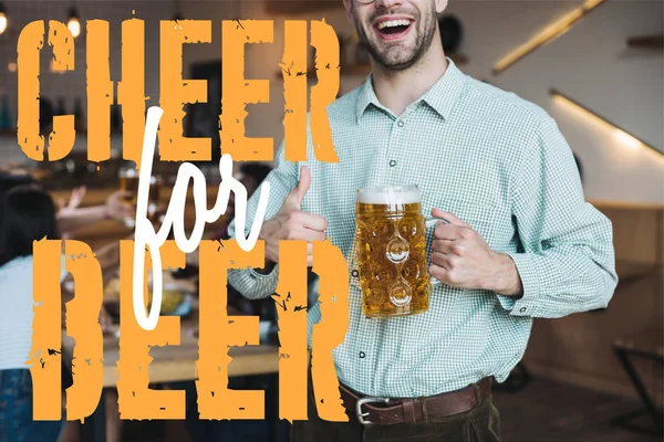 Vista recortada de sonriente joven sosteniendo taza de cerveza lager y mostrando el pulgar hacia arriba cerca de alegría por la ilustración de la cerveza - foto de stock