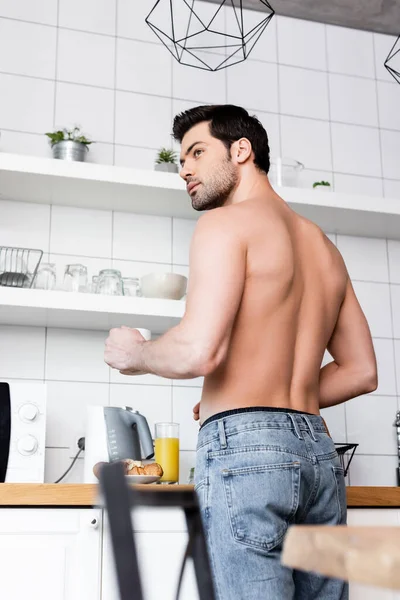 Hombre sin camisa guapo sosteniendo taza de café en la cocina con desayuno - foto de stock