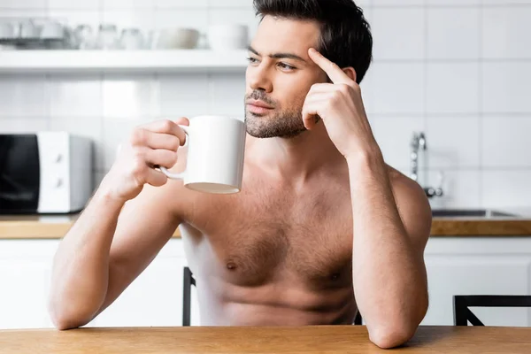 Nachdenklicher, hemdsloser Mann, der morgens eine Tasse Kaffee in der Küche hält — Stockfoto