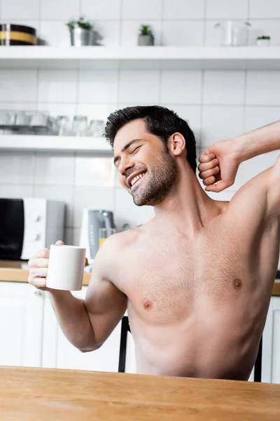 Happy shirtless man stretching and holding cup of coffee on kitchen in morning — Stock Photo