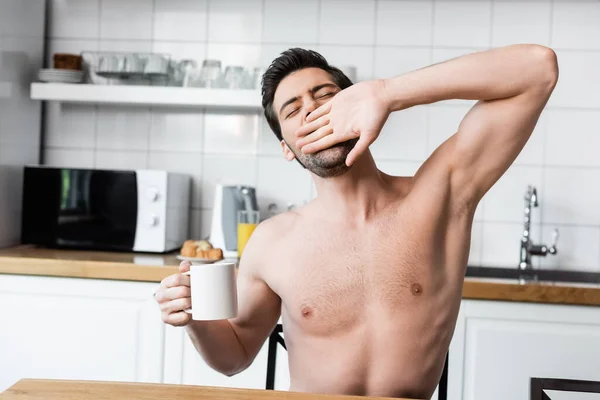 Uomo senza maglietta che tiene in mano una tazza di caffè mentre sbadiglia in cucina al mattino — Foto stock