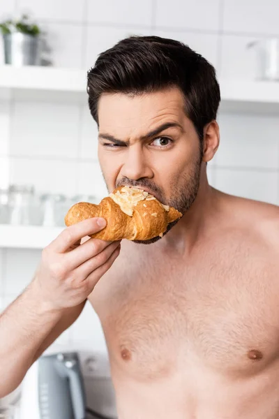 Шокований без сорочки чоловік їсть круасан на кухні вранці — стокове фото
