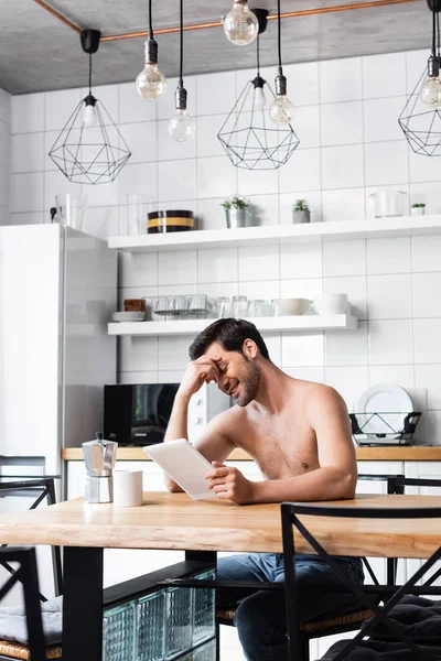 Веселый мужчина без рубашки с помощью цифрового планшета на кухне с кофейником и чашкой — стоковое фото