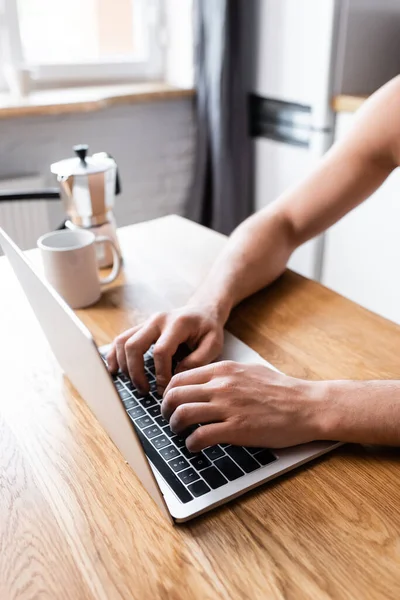 Обрезанный вид мужчины фрилансера, работающего на ноутбуке на кухне с кофейником и чашкой — стоковое фото