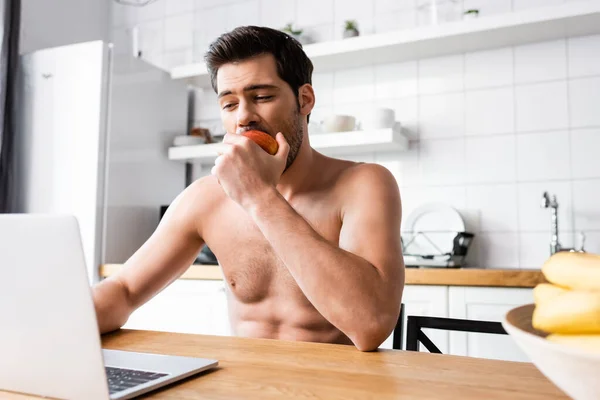 Красивый фрилансер без рубашки ест яблоко во время работы над ноутбуком на кухне — стоковое фото