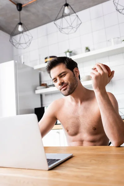 Усміхнений фрілансер без сорочки їсть яблуко під час роботи на ноутбуці на кухні — стокове фото