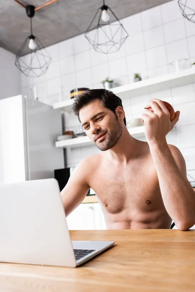 Sourire torse nu pigiste manger des pommes tout en travaillant sur un ordinateur portable sur la cuisine — Photo de stock