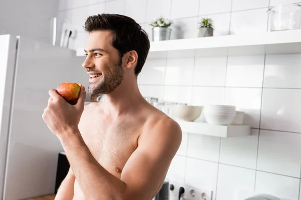 Веселий без сорочки чоловік їсть яблуко на кухні вранці — стокове фото