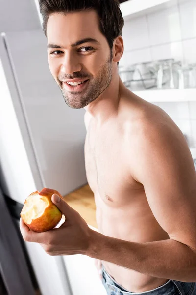 Feliz hombre sin camisa comiendo manzana en la cocina en la mañana - foto de stock