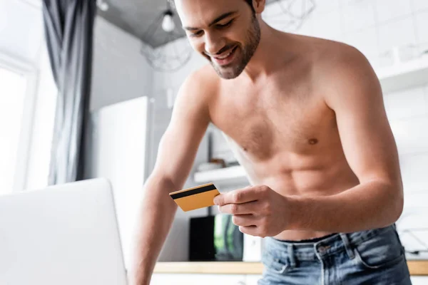 Cheerful shirtless man shopping online with credit card and laptop on kitchen — Stock Photo