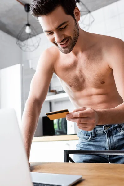 Sonriente hombre sin camisa de compras en línea con tarjeta de crédito y portátil en la cocina - foto de stock