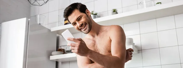 Смеющийся мужчина без рубашки с помощью смартфона на кухне дома, панорамный урожай — стоковое фото