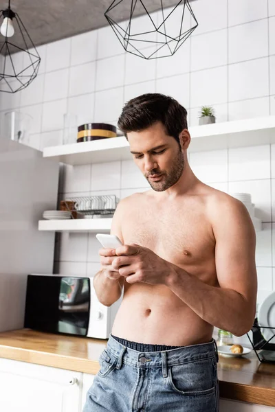 Красивый мужчина без рубашки с помощью смартфона на кухне дома — стоковое фото