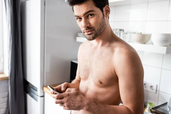 Сексуальный мужчина без рубашки с помощью смартфона на кухне дома — стоковое фото