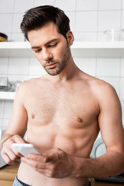 Schöner Mann ohne Hemd benutzt Smartphone in der Küche zu Hause — Stockfoto