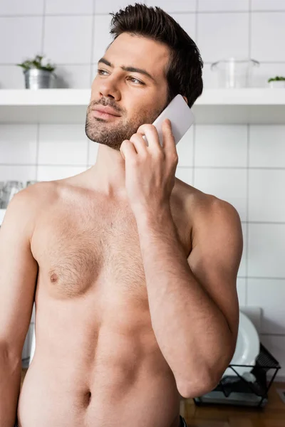 Homem sem camisa pensativo falando no celular na cozinha em casa — Fotografia de Stock