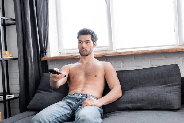 Bonito sem camisa homem segurando controle remoto enquanto assistindo tv em casa — Stock Photo