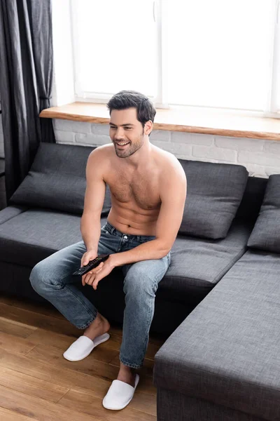 Sexy sorrindo homem segurando controle remoto enquanto assistindo tv em casa — Fotografia de Stock