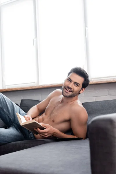 Guapo feliz sin camisa hombre leyendo libro en sofá - foto de stock