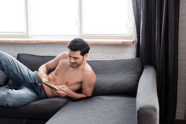 Bell'uomo sorridente senza maglietta che legge il libro sul divano — Foto stock