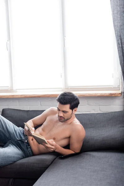 Красивый сексуальный мужчина без рубашки читает книгу на диване — стоковое фото
