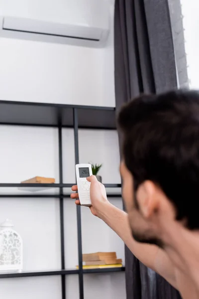 Young man with remote controller adjusting temperature of air conditioner at home, selective focus — Stock Photo