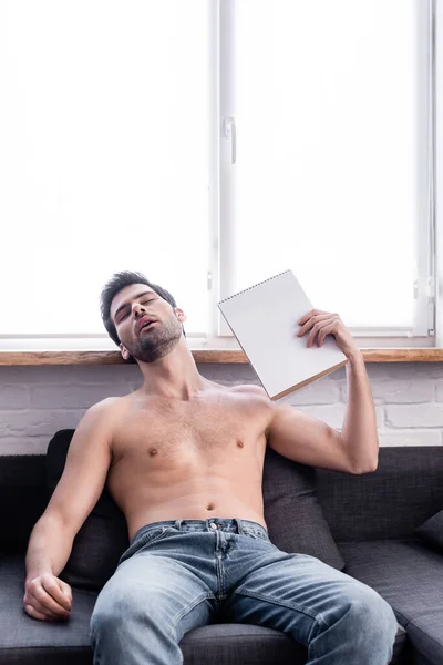 Hombre sin camisa saludando con bloc de notas como ventilador mientras sufre de calor en casa - foto de stock